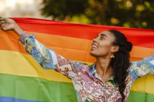 A young person proudly holding a rainbow flag and smiling outdoors, symbolizing LGBTQ+ pride, hope, and the importance of supportive mental health and addiction for dual diagnosis treatment.