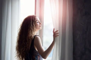 Woman standing by a window with soft sunlight, gently opening the curtains and breathing in fresh air.